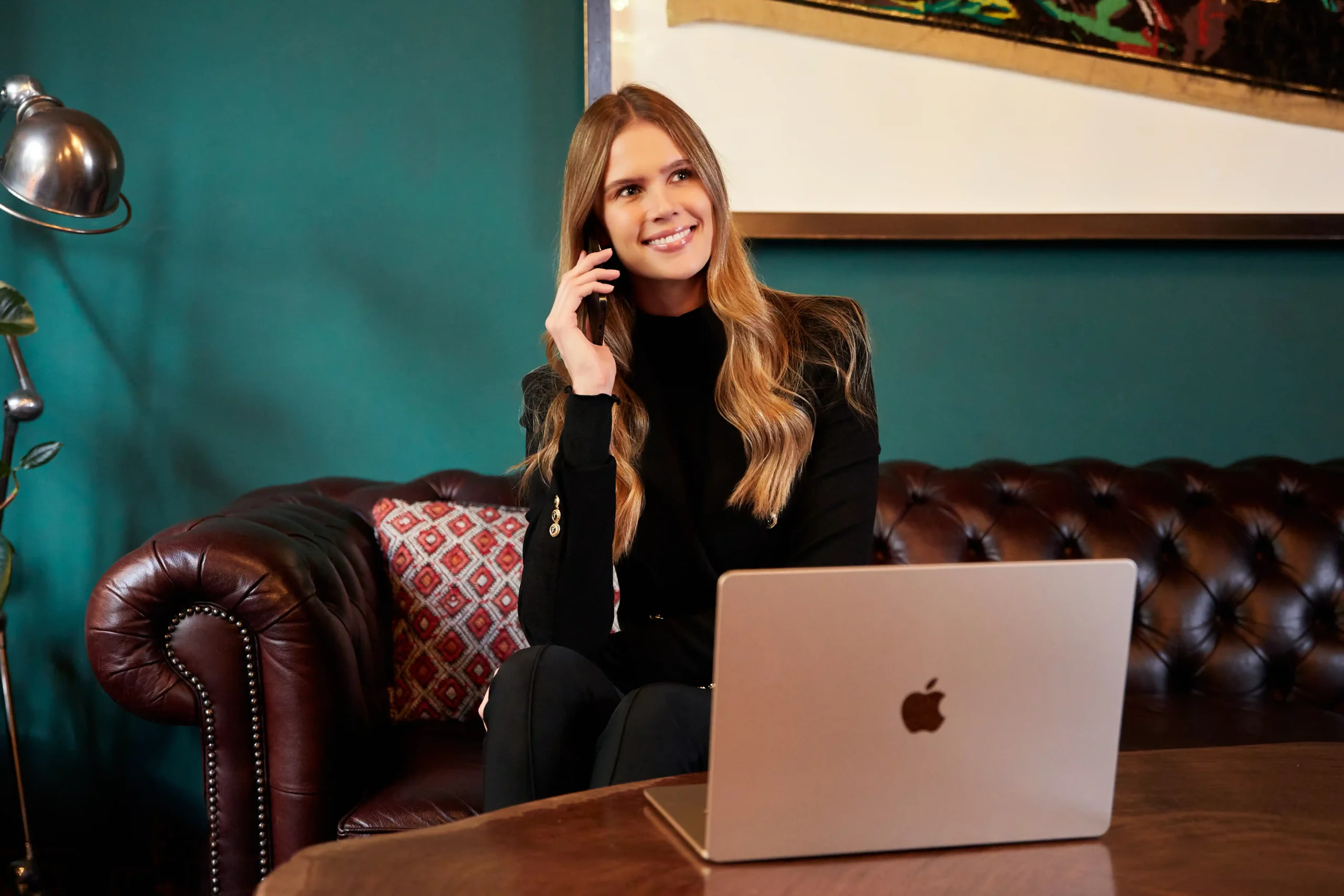 Smiling woman sitting on leather sofa talking on phone with laptop.