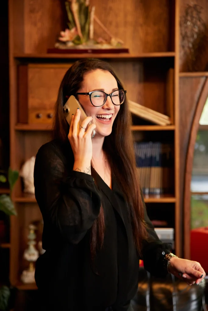 Smiling woman with glasses talking on phone in cozy office setting.